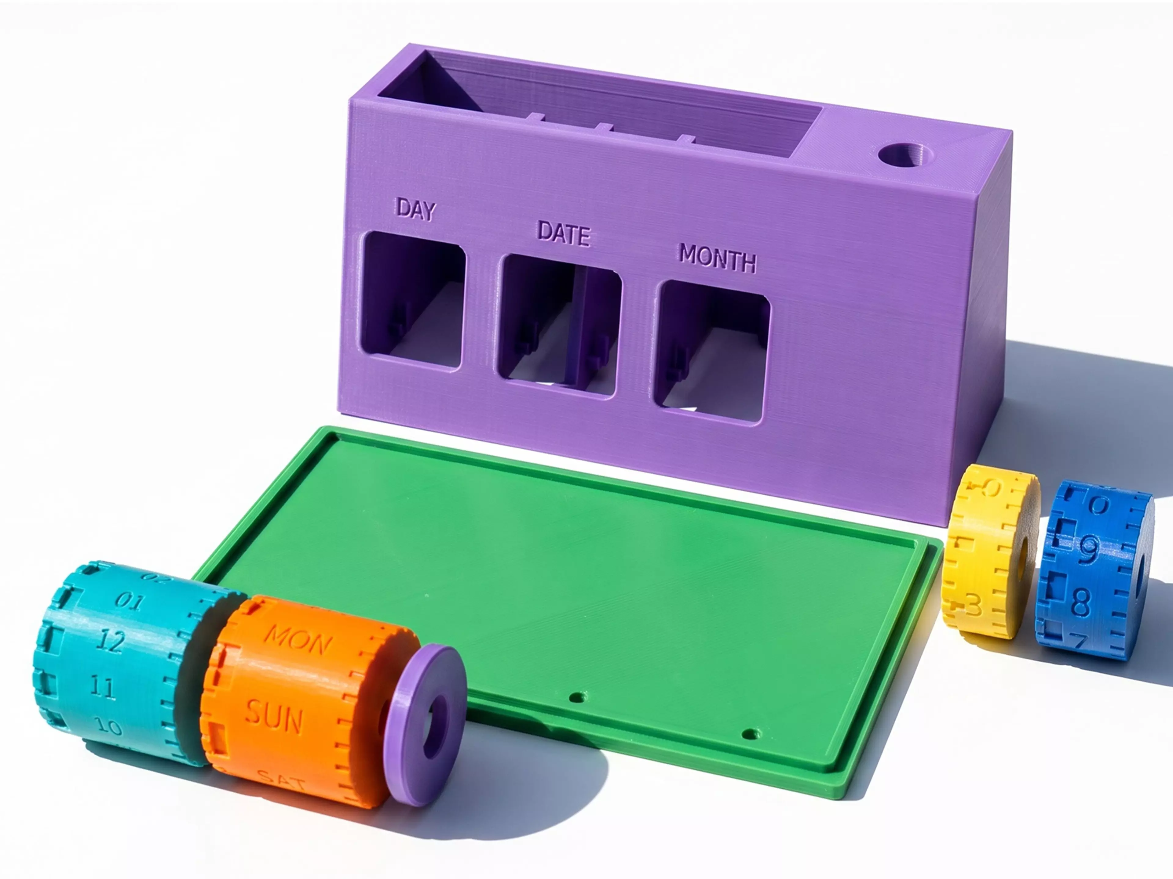 A mechanical desk organizer/perpetual calendar: a matte-white wedge-shaped body with three independently spinning charcoal drums (weekday / day / month, viewed through front windows), a teal accent dial ring on the right end, a rear pen slot, and an open storage tray — no electronics.