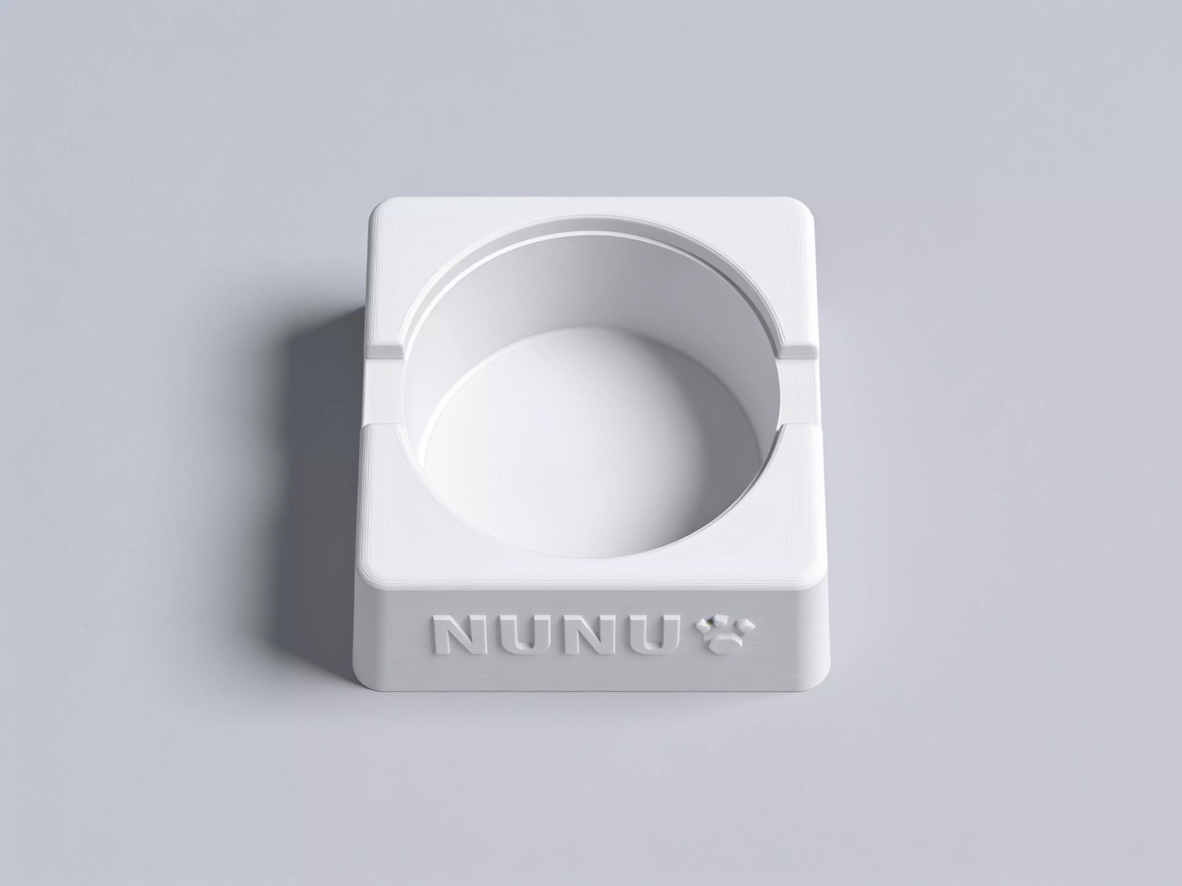 A single-piece, Scandinavian-minimal squircle stand (200 × 200 × 76 mm) that a standard 1-quart stainless bowl drops into from the top and hangs by its rolled rim on an internal support lip, with an anti-spill raised rim, two finger scallops for bowl removal, "NUNU" + paw icon raised on the front…