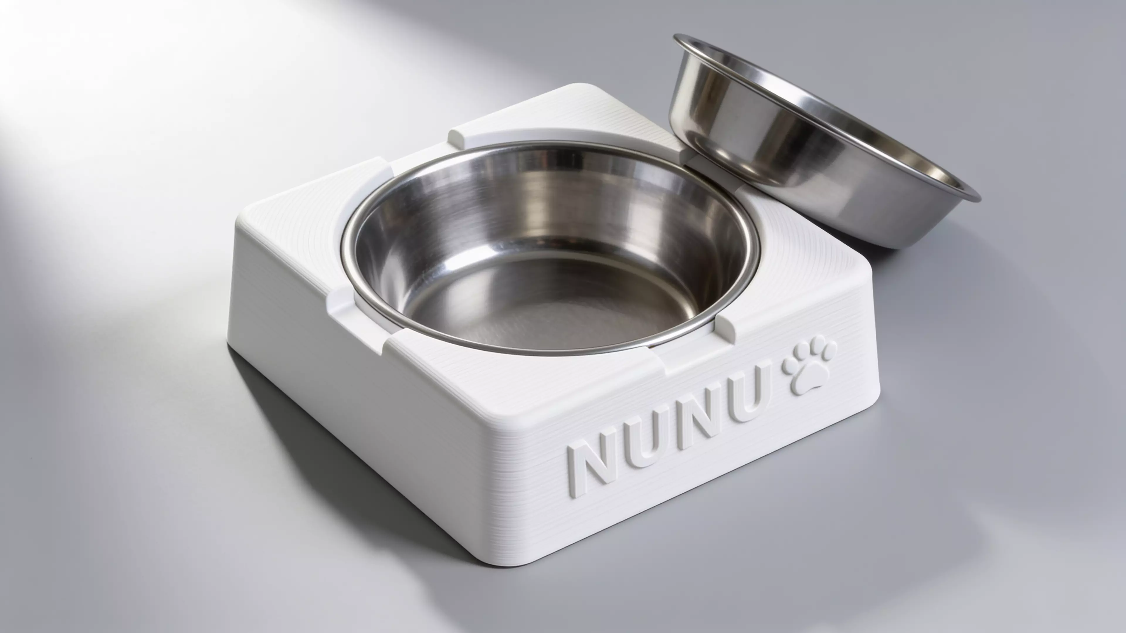 A single-piece, Scandinavian-minimal squircle stand (200 × 200 × 76 mm) that a standard 1-quart stainless bowl drops into from the top and hangs by its rolled rim on an internal support lip, with an anti-spill raised rim, two finger scallops for bowl removal, "NUNU" + paw icon raised on the front…