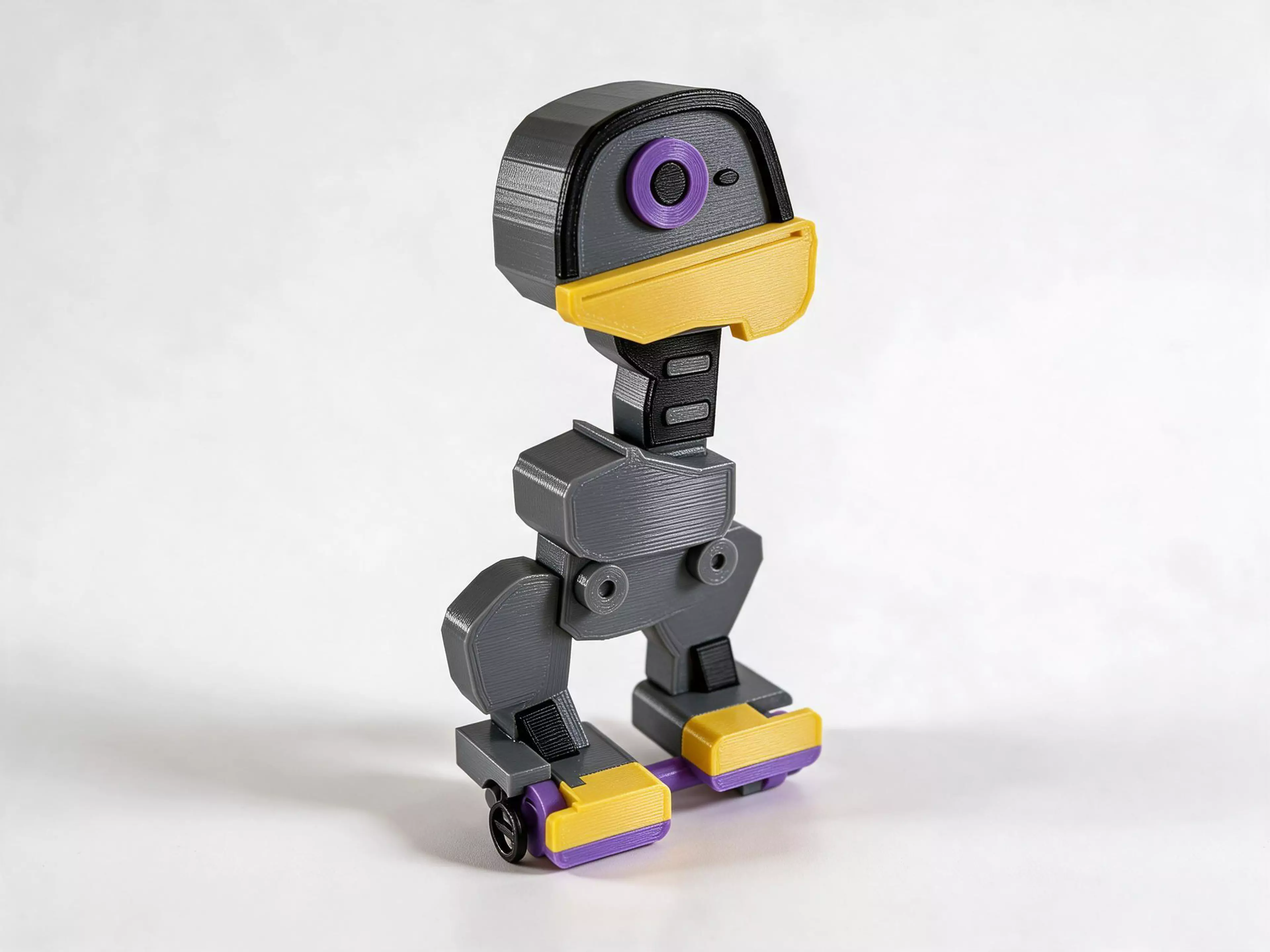 A grey robot duck with a yellow bill, purple details and a side winding knob. A replaceable silicone cord stores the twist that drives its small wheels.