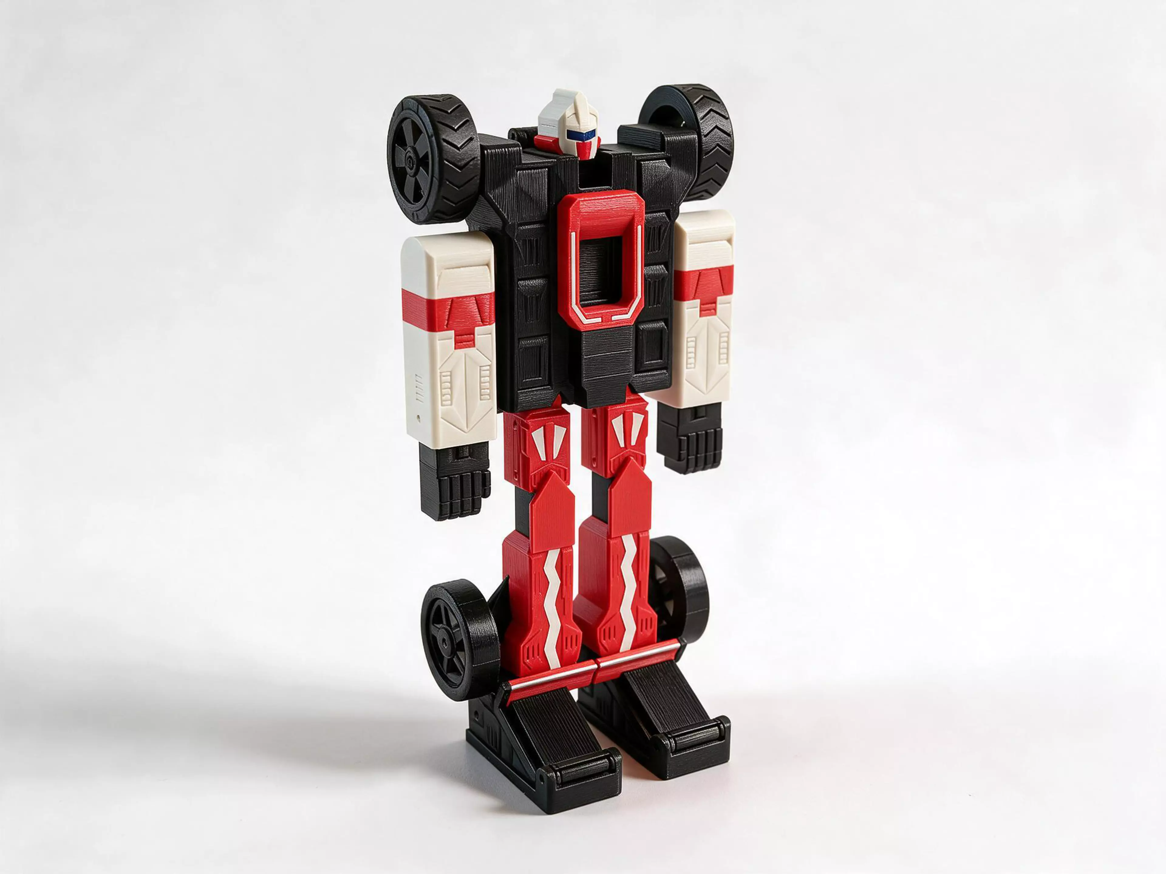 A 250 mm heroic robot in layered red, white and dark gray armour that changes by hand, with no part removed, into a 337 mm open-wheel Formula racing car on four free-turning wheels. Its nose tip now slides into the nose so the car sits shorter, the slick rear wheels ride low on the shin cuffs beside the boots, and both wheel pairs are larger for more ground clearance. Every hinge grips its pin in a split bore, so the nose, fists and rear-wing halves hold their angle. Wedge boots carry the two rear-wing halves on toe hinges, and a red halo rings the cockpit on its chest and on top of the car. The printed bodies carry press-fit colour pieces and join on 3 mm steel dowels: no glue and no supports.